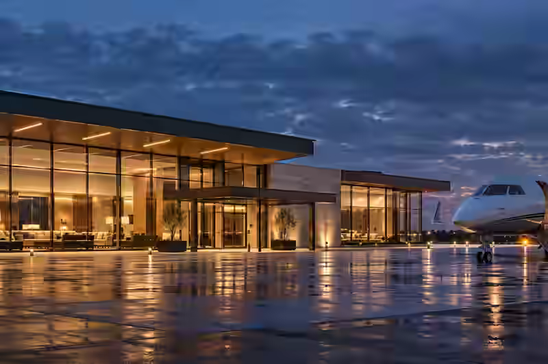 Conceptual glass private aviation terminal glowing at blue hour beside a generic business jet on wet tarmac